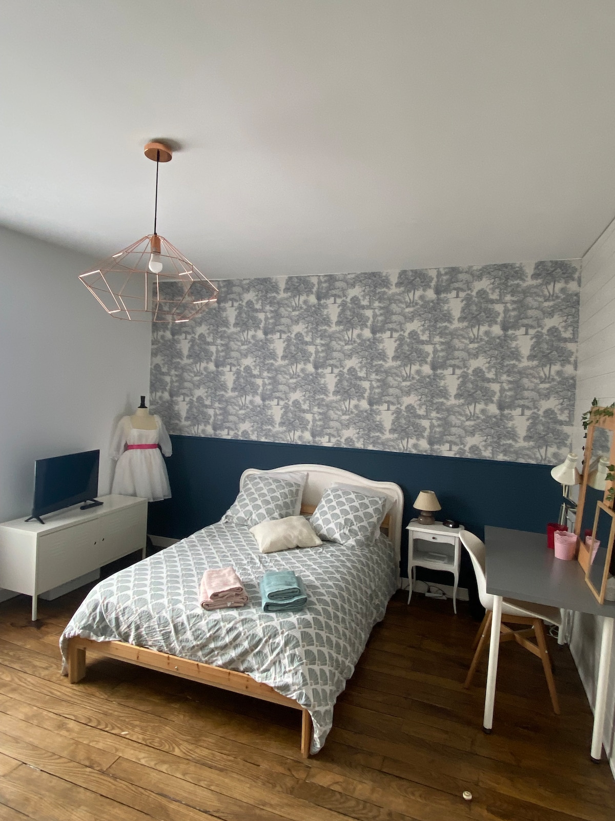 A well-appointed bedroom is presented, featuring a spacious bed with a patterned duvet and decorative pillows. A small desk is positioned beside the bed, and a television is visible on a low cabinet. The decor includes a light fixture and a dress form in the corner.