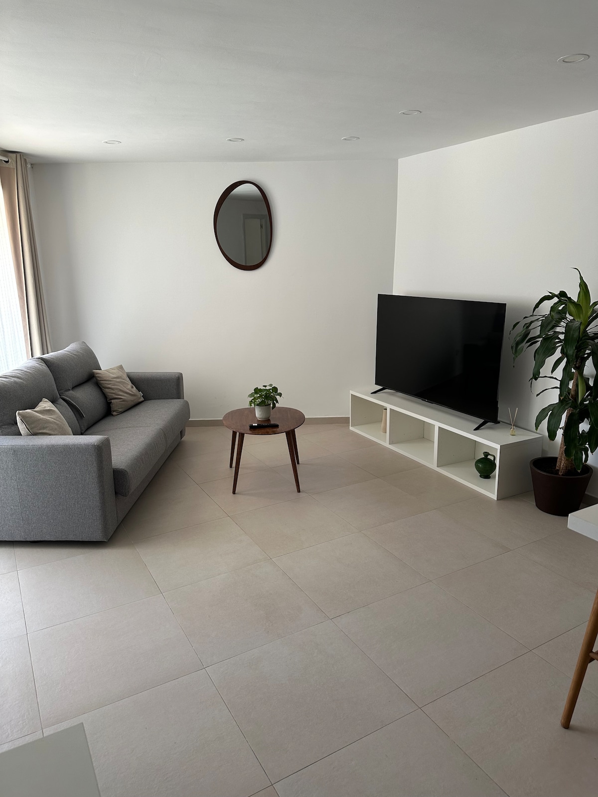 A modern living space is depicted, featuring a gray sofa accompanied by decorative pillows. A circular mirror adorns the wall above a small plant on a wooden side table. A large television is placed on a minimalist white console, while a tall green plant adds a touch of nature.
