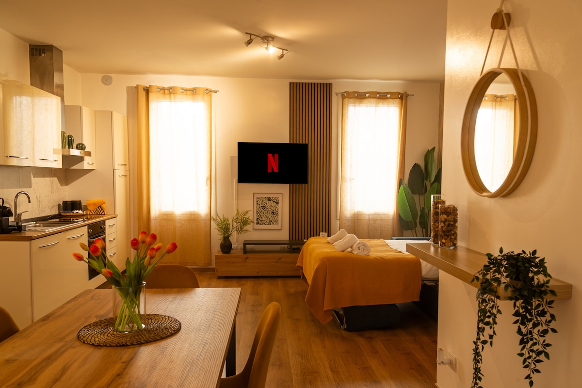 A bright living area features a Smart TV mounted on the wall. A spacious sofa bed is arranged next to large windows with light curtains. A dining table with chairs sits nearby, adorned with a vase of tulips. A circular mirror adds depth to the space.