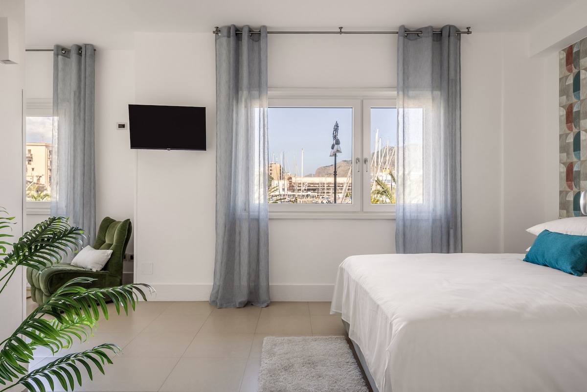 A bright and airy bedroom is furnished with a spacious bed and a cozy green armchair. Natural light filters through sheer curtains, illuminating the sleek design. A wall-mounted Smart TV adds convenience, while a view of the marina is framed by the window.
