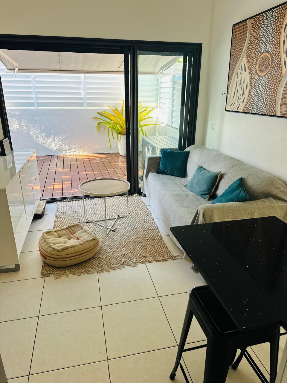 The living area is designed with a light-colored sofa and a round coffee table, situated on a textured rug. Large sliding glass doors lead to an outdoor deck area, where potted plants are visible. A dining table with black metal legs complements the modern decor.