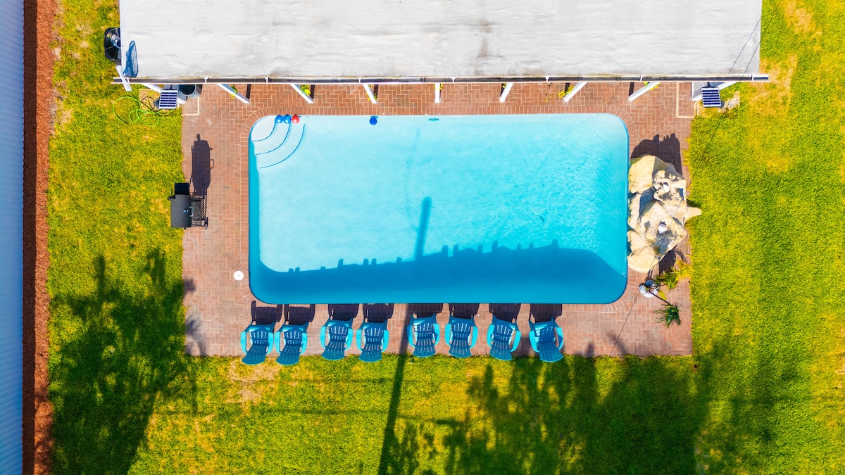 An aerial view showcases a spacious swimming pool surrounded by neat, green lawns. Several lounge chairs line the poolside, providing ample seating space. The area is set under a covered patio, offering a shaded spot for relaxation.
