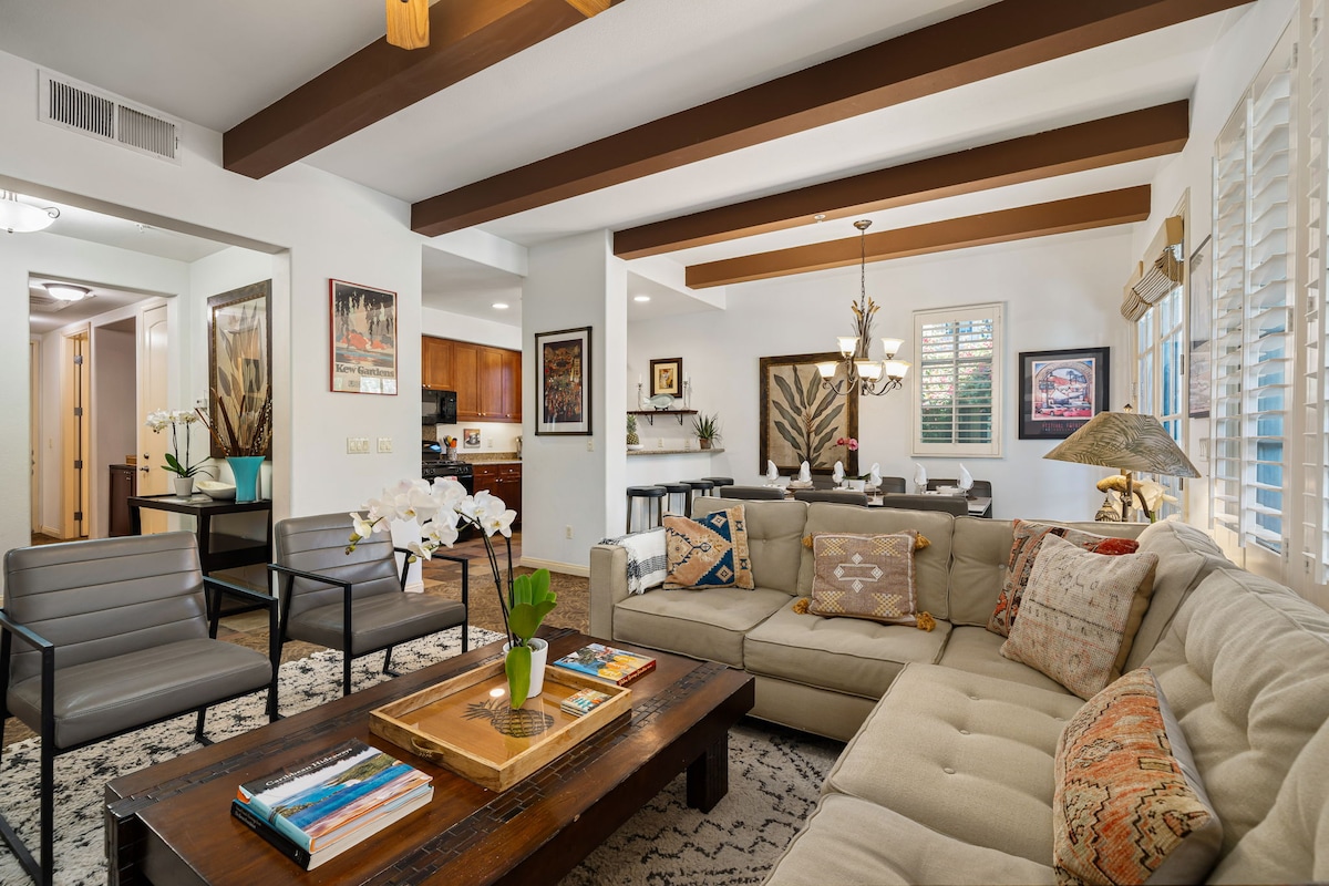 A welcoming living area features a spacious sectional sofa with decorative pillows, centered around a coffee table. Wood beams line the ceiling, and light comes through shutters on the windows. A dining area and kitchen are visible in the background, accentuating the open layout.