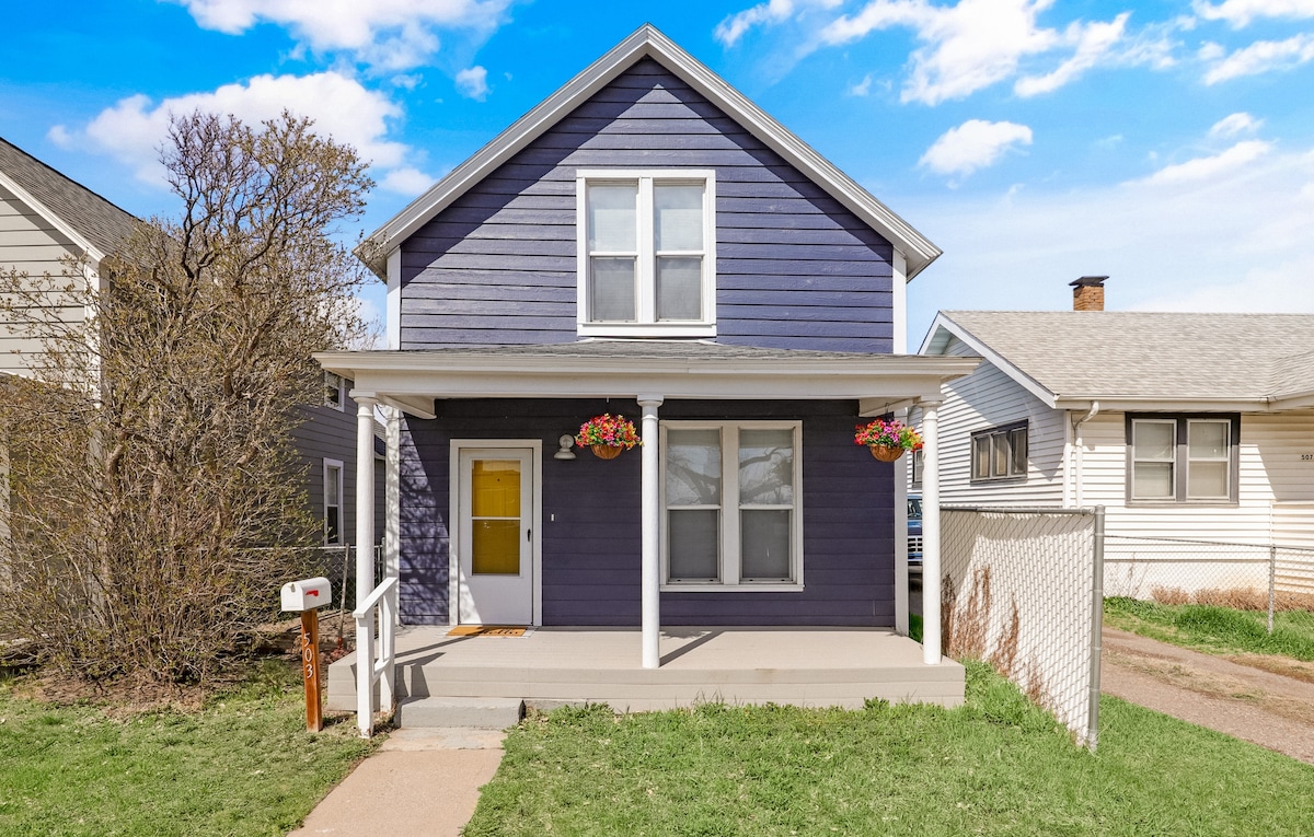 The charming blue house features a welcoming front porch with a white railing. Flower baskets are hung on the porch pillars, adding a pop of color. Large windows allow natural light to flow in, while the surrounding greenery contributes to the inviting exterior.