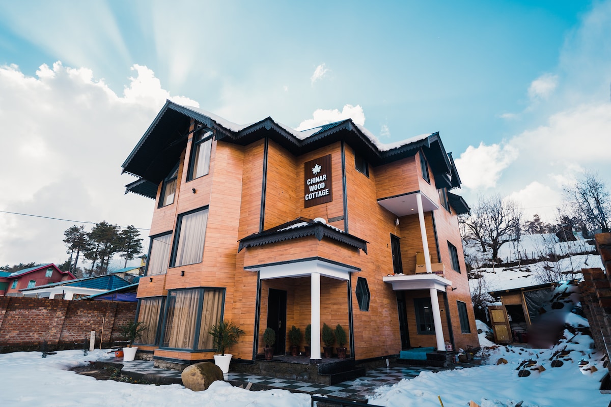 A wooden cottage is presented with a modern design, featuring multiple levels and large windows that reflect the surrounding landscape. Snow covers the ground, and lush greenery is visible nearby, creating a serene environment. The cottage's structure displays a blend of natural materials and contemporary architecture.