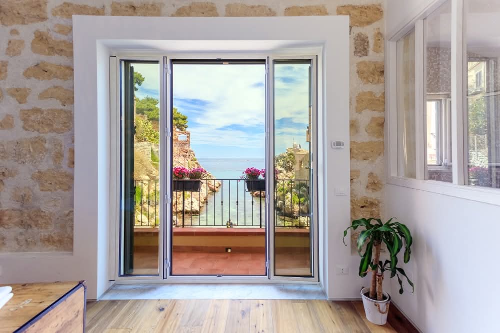 Large glass doors open to a balcony with a view of the serene caletta. Potted plants and vibrant flowers add color to the balcony railing, framed by natural stone walls. The indoor space features wooden flooring, contributing to a warm and inviting environment.