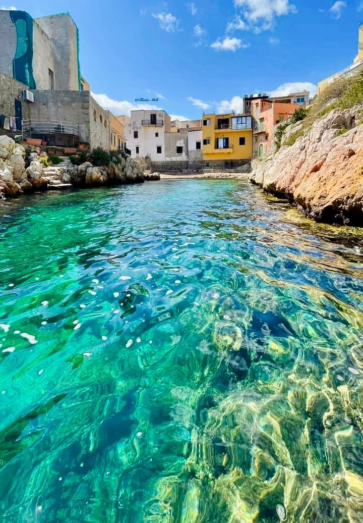 A serene coastal scene showcases crystal-clear turquoise water, gently reflecting the colors of surrounding buildings. The rocky shoreline leads to inviting structures with vibrant hues, creating a harmonious view that captures the essence of seaside living.