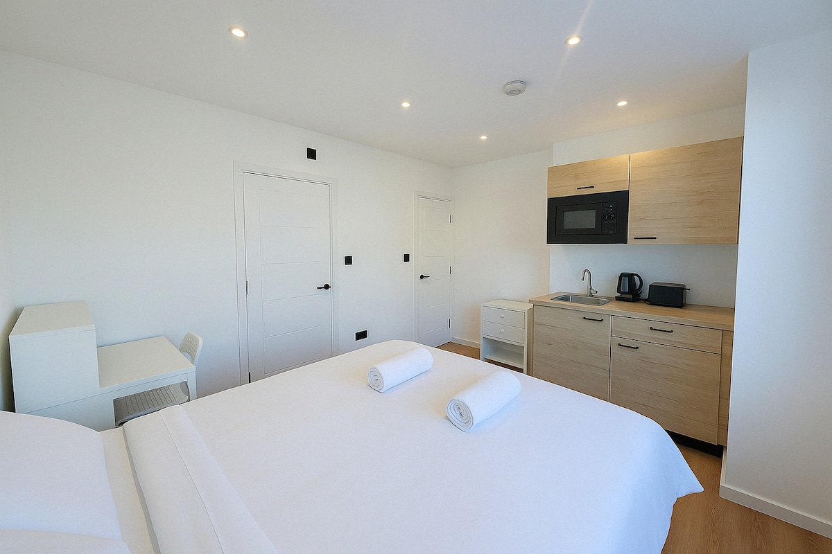 A bright studio space features a spacious bed with neatly arranged towels. Light wooden cabinetry and a kitchenette area include a microwave, sink, and kettle. A small dining table and chair are positioned next to the bed, while white walls create an airy feel in the room.