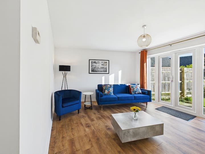 A spacious, modern living room with a three-seater sofa, two matching tub chairs, and a sofa bed option. Enjoy a 50-inch TV with Sky TV and Netflix. The room opens to a large garden, perfect for sunny days.
