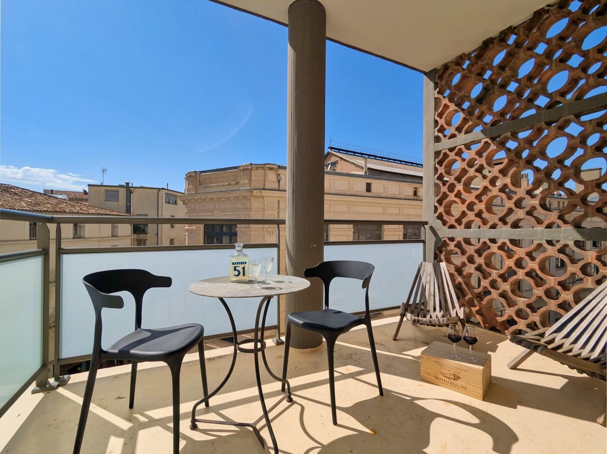 The terrace features a small round table accompanied by two black chairs. A bottle of water and two glasses sit on the table. Folding sun loungers are visible in the background, with a decorative wall enhancing the space. Bright sunlight illuminates the area.