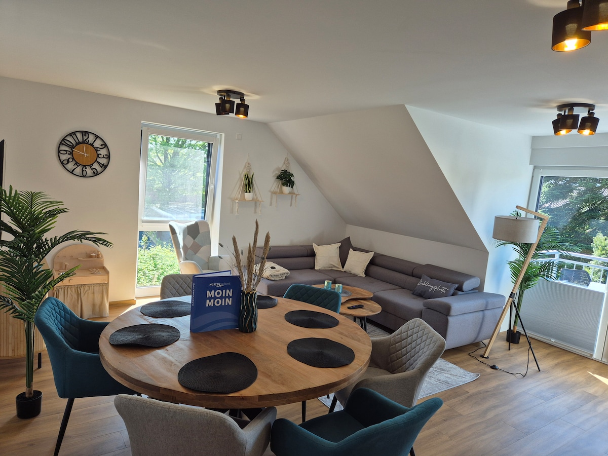 A modern living area features a circular wooden dining table surrounded by upholstered chairs, alongside a spacious gray sectional sofa. Large windows allow natural light to fill the room, complemented by decorative plants and a stylish floor lamp. A welcoming atmosphere is created for relaxation and dining.