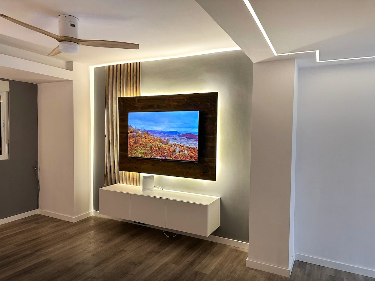 A modern living area features a wall-mounted television framed by a wooden accent. Soft lighting is integrated along the edges, enhancing the overall ambiance. A low, white media console sits beneath the television, complementing the light wooden flooring.