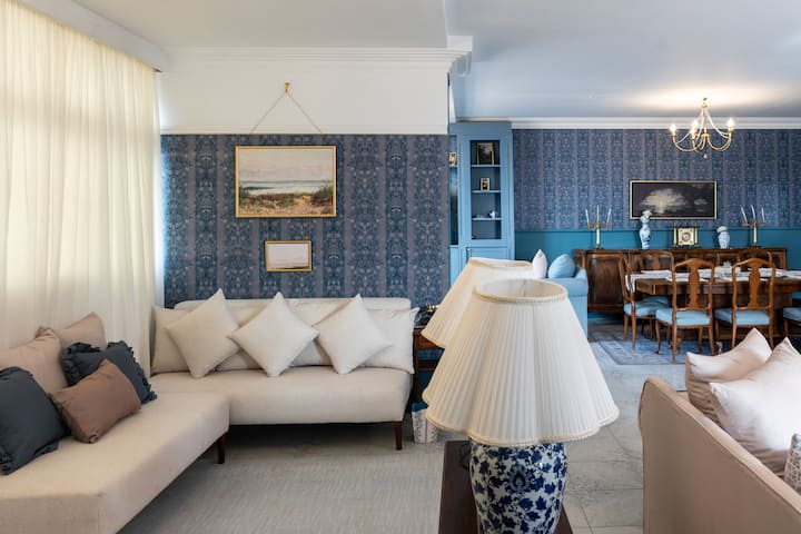 Este elegante espacio de estar y comedor cuenta con una decoración clásica, sofás de color crema adornados con lujosas almohadas y muebles de madera. Una lámpara de araña ilumina el comedor, mientras que las lámparas decorativas y las pinturas enmarcadas mejoran el ambiente acogedor.