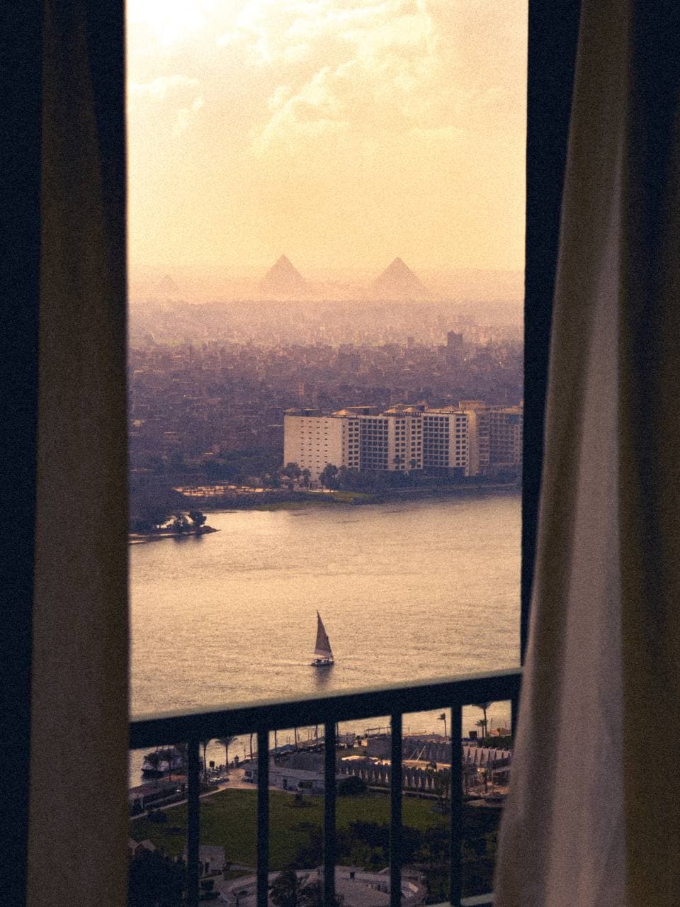 An inviting view from a window shows the Nile River and distant Great Pyramids under a soft, hazy sky. A sailboat gently glides across the water, while city buildings blend with the natural landscape surrounding the river.