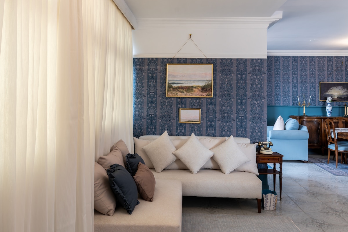 A stylish living area features a light-colored sofa adorned with several cushions, complemented by a decorative side table. The walls are decorated with elegant patterns and framed artwork, while sheer curtains allow natural light to softly illuminate the space.