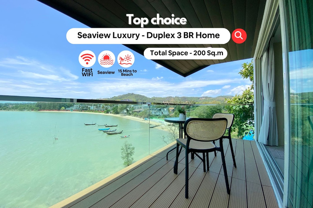 A private balcony offers a direct view of the shimmering sea, featuring a small table and two chairs. The surrounding area is adorned with greenery and hills in the distance. The calm waters reflect the sunny sky, creating a serene environment.