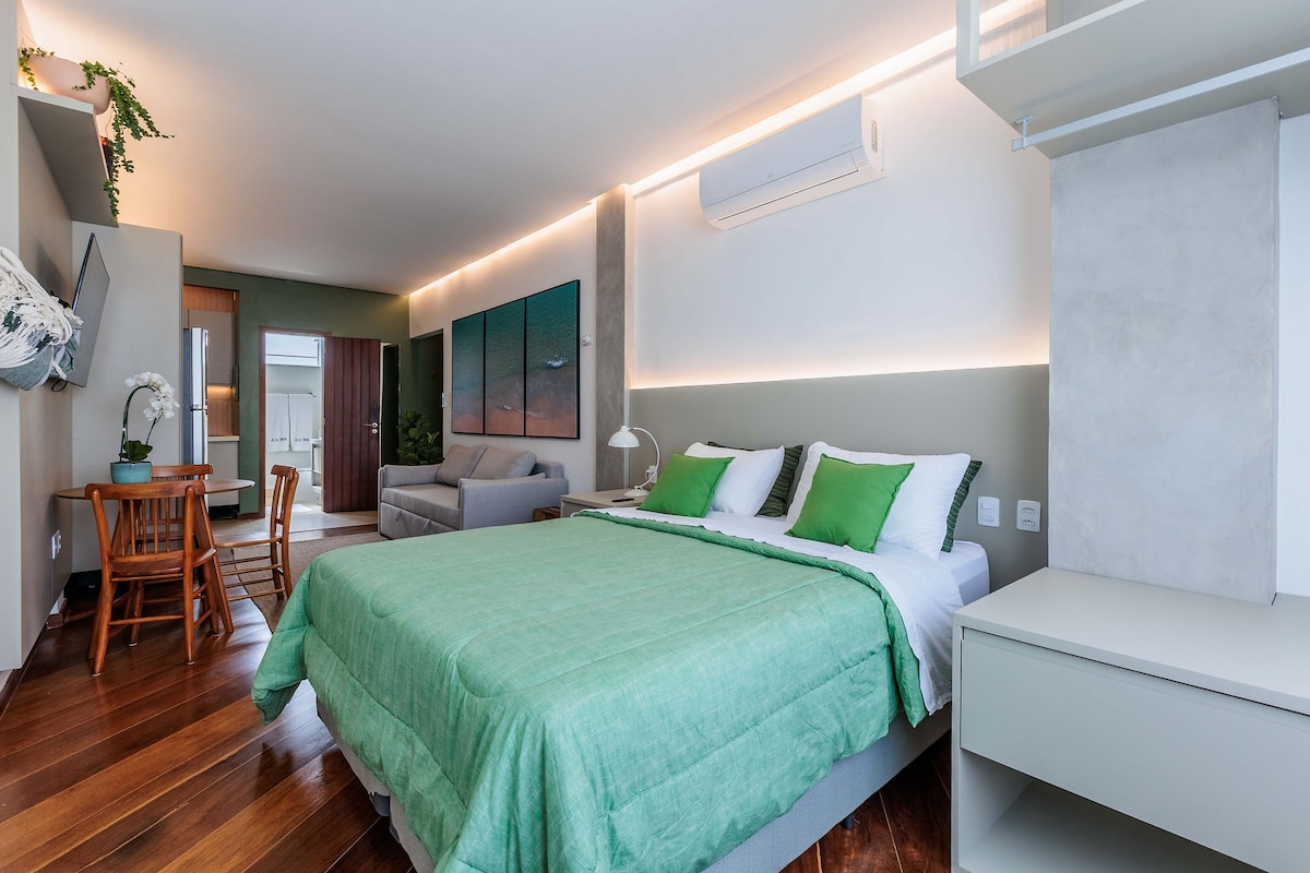 A spacious studio is presented, featuring a large bed with a textured green blanket. Soft lighting illuminates the room, highlighting a comfortable seating area with a gray sofa. A dining set for two is visible, along with a light-colored cabinet and wall-mounted television.