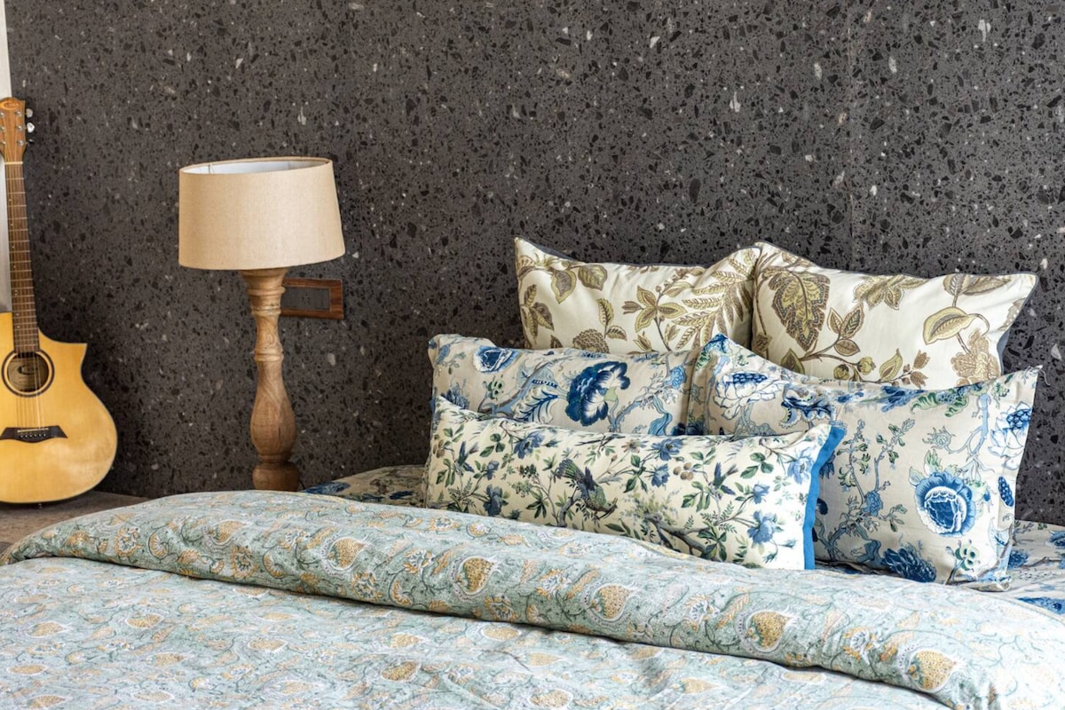 A spacious bed is adorned with an assortment of decorative pillows in floral patterns. A soft blanket with intricate designs lies at the foot of the bed. A wooden bedside lamp provides warm lighting, while a guitar leans against the textured wall.