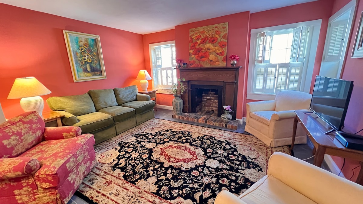 The living area features a cozy seating arrangement with a green sofa, a pink armchair, and a white chair. A brick fireplace is surrounded by decorative items. Natural light enters through the windows, which are adorned with shutters, highlighting the vibrant orange walls.