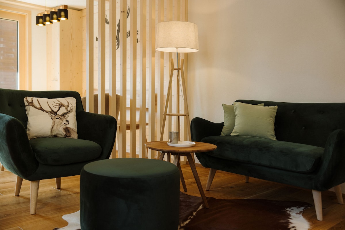 A seating area is furnished with two plush green sofas, a round ottoman, and a wooden coffee table. A floor lamp provides soft lighting. Natural wood accents and light-colored walls create a cozy and inviting environment.