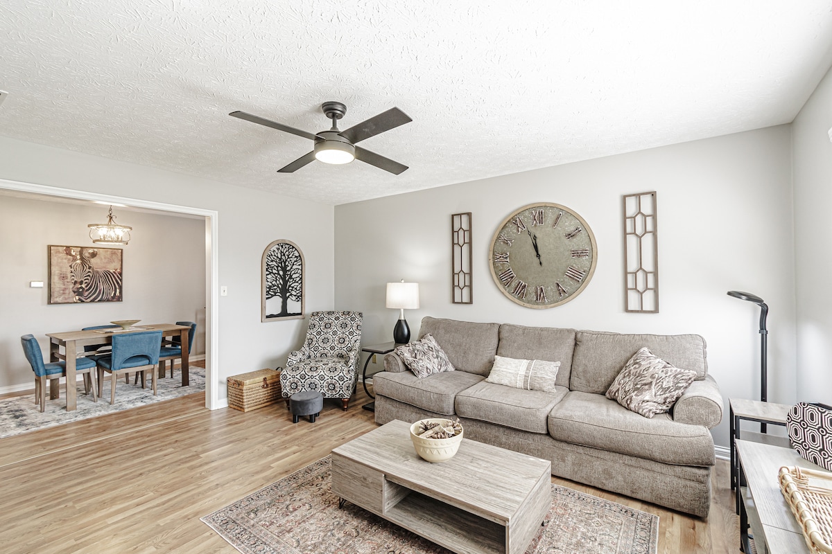 A spacious living area features a comfortable sofa with decorative cushions and a textured area rug. A ceiling fan enhances air circulation while a large wall clock adds a focal point. A dining area with blue chairs is visible through an open entryway, enhancing connectivity.