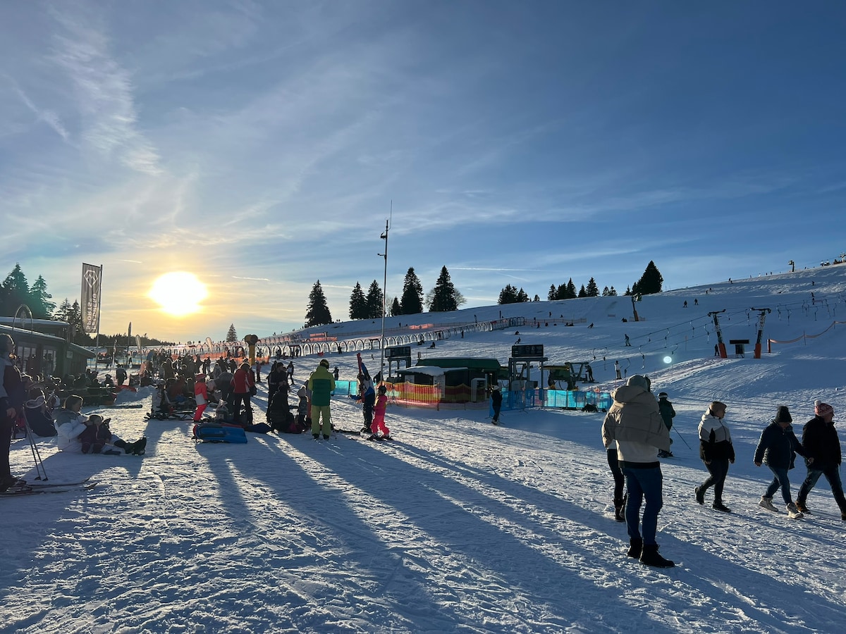 A snowy landscape is filled with skiers and visitors enjoying the winter sun. The slopes are alive with activity, while a ski lift can be seen in the distance. Shadows stretch across the snow, reflecting the low winter sun setting in the background.
