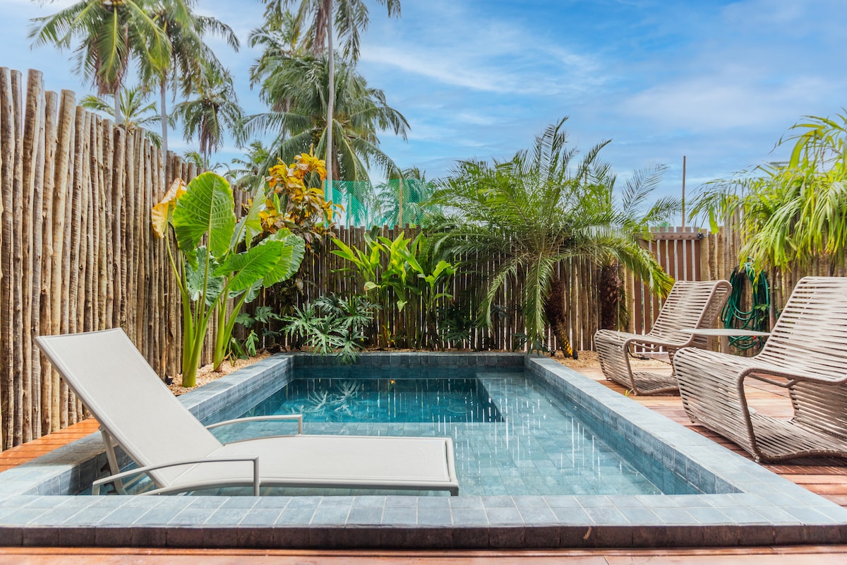 A private pool area is surrounded by lush tropical plants and palm trees. A reclined lounge chair is positioned beside the rectangular pool, which reflects the clear blue sky. Wooden decking frames the pool, enhancing the serene atmosphere.