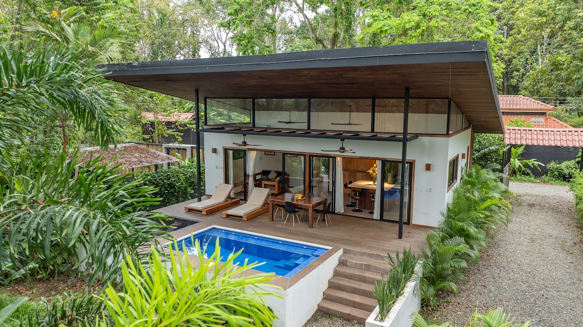 A modern, one-story house is surrounded by lush greenery, featuring a private pool on a spacious wooden deck. Large glass sliding doors provide views into the inviting interior, while two lounge chairs sit beside the pool, perfect for relaxation in a tranquil setting.
