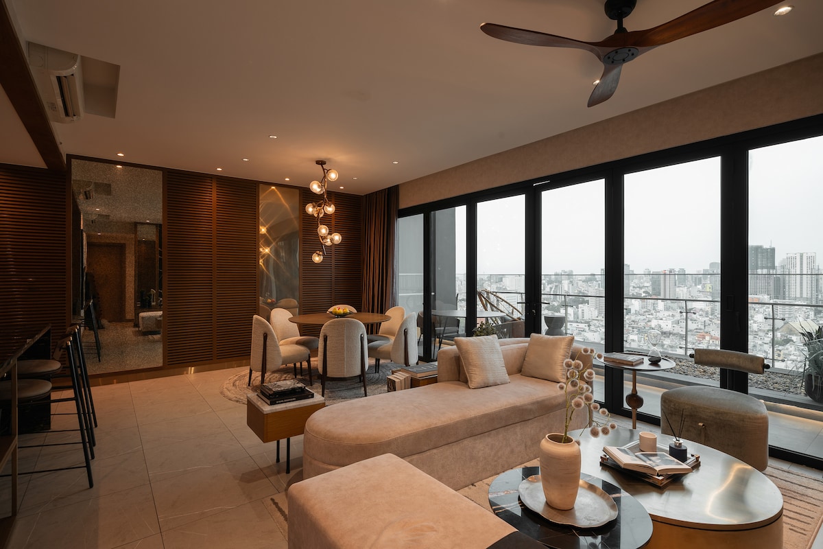 A spacious living area features large floor-to-ceiling windows that provide expansive views of the city skyline. Modern furniture includes a light-colored sectional sofa and stylish chairs around a round dining table. A ceiling fan adds comfort, while decorative elements enhance the contemporary design.
