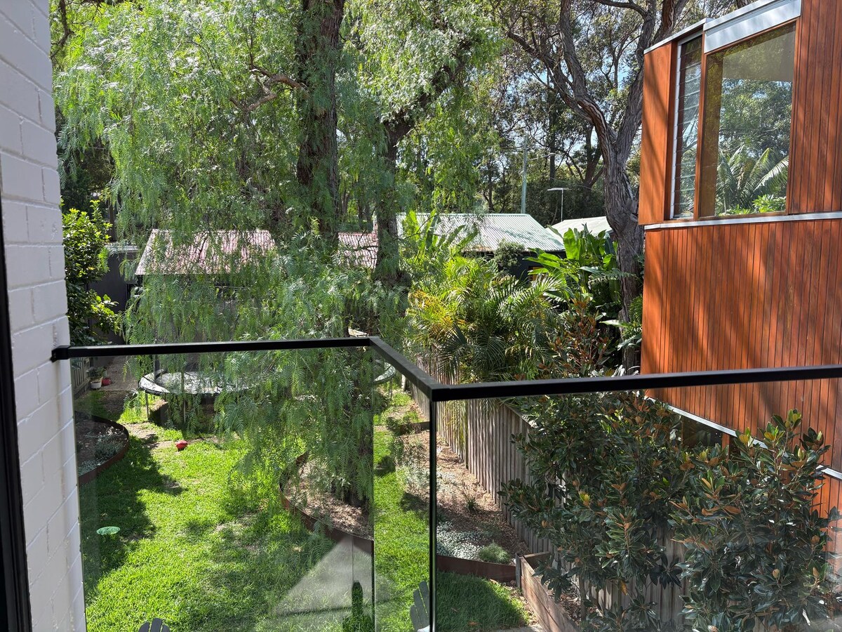 A private balcony offers a view of a lush garden filled with greenery and trees. The area features a well-maintained lawn and nearby structures, enhancing the serene outdoor ambiance. Natural light filters through the foliage, creating a tranquil setting.