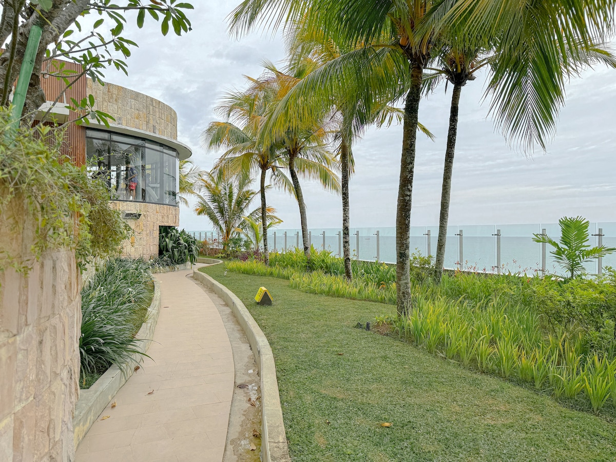 A pathway meanders through lush greenery, bordered by tall palm trees. The ocean is visible in the distance, providing a serene backdrop. The gently curved building structure features large windows, inviting natural light. Well-maintained landscaping enhances the peaceful outdoor setting.