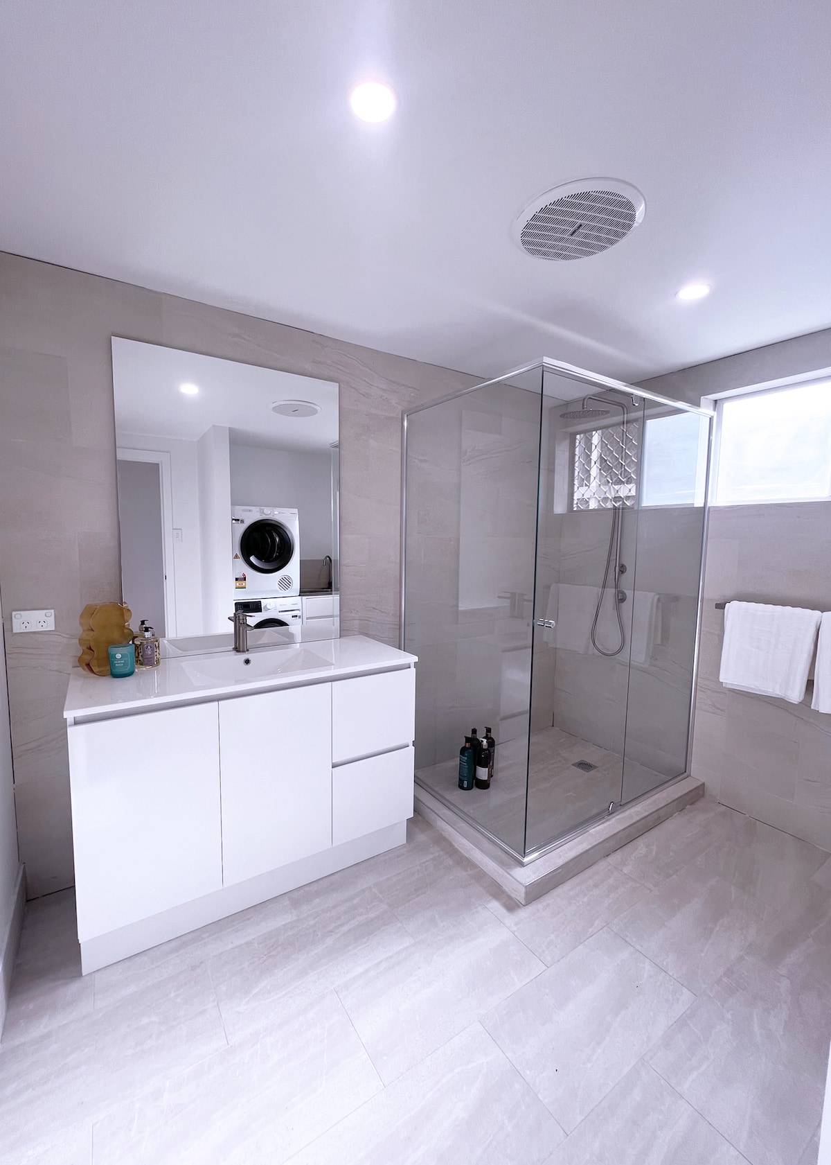 A modern bathroom features a spacious shower with glass enclosure and chrome fixtures. A sleek vanity with a large mirror is situated beside the shower. Natural light enters through a window, illuminating the light-colored tiles and soft tones throughout the space.