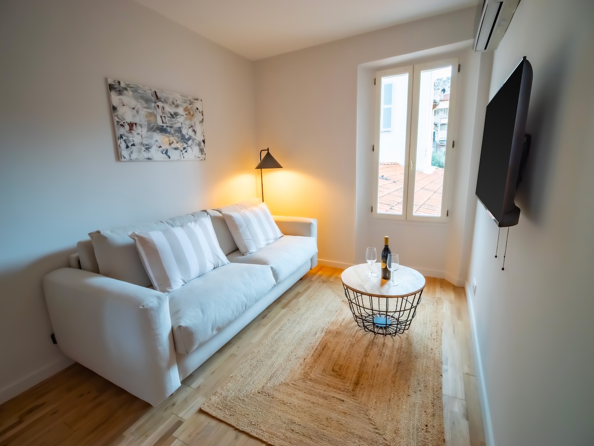 A bright living area is shown, featuring a light-colored sofa adorned with neutral pillows. A round coffee table is positioned center stage, accompanied by a bottle of wine. Natural light filters softly through a window, creating a calm atmosphere in the space.
