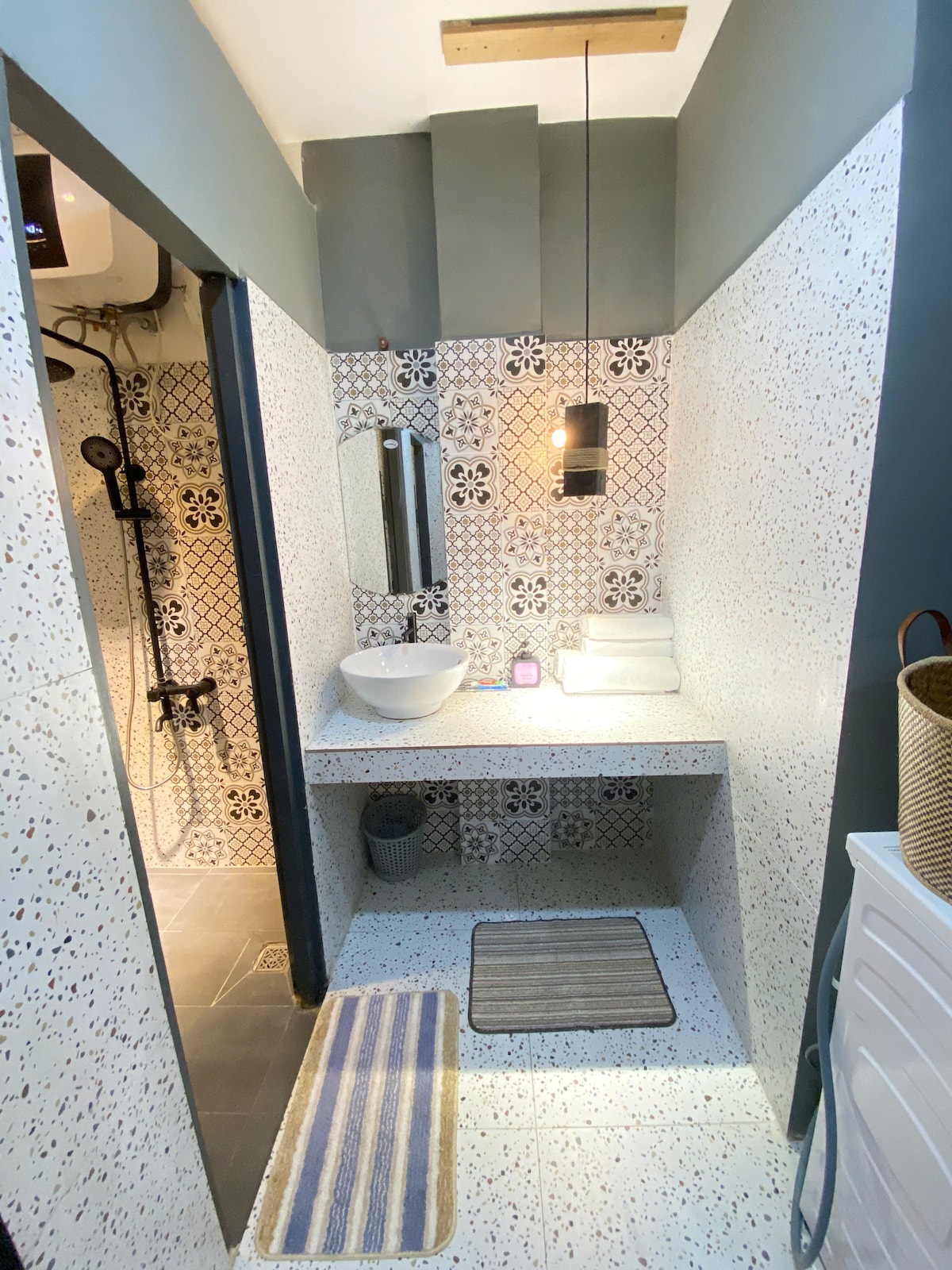 The bathroom features patterned tiles complemented by a modern sink and mirror. Soft lighting is provided by a hanging lamp. Fresh towels are neatly arranged on a countertop, and the space includes a small bin and floor mats to enhance comfort.