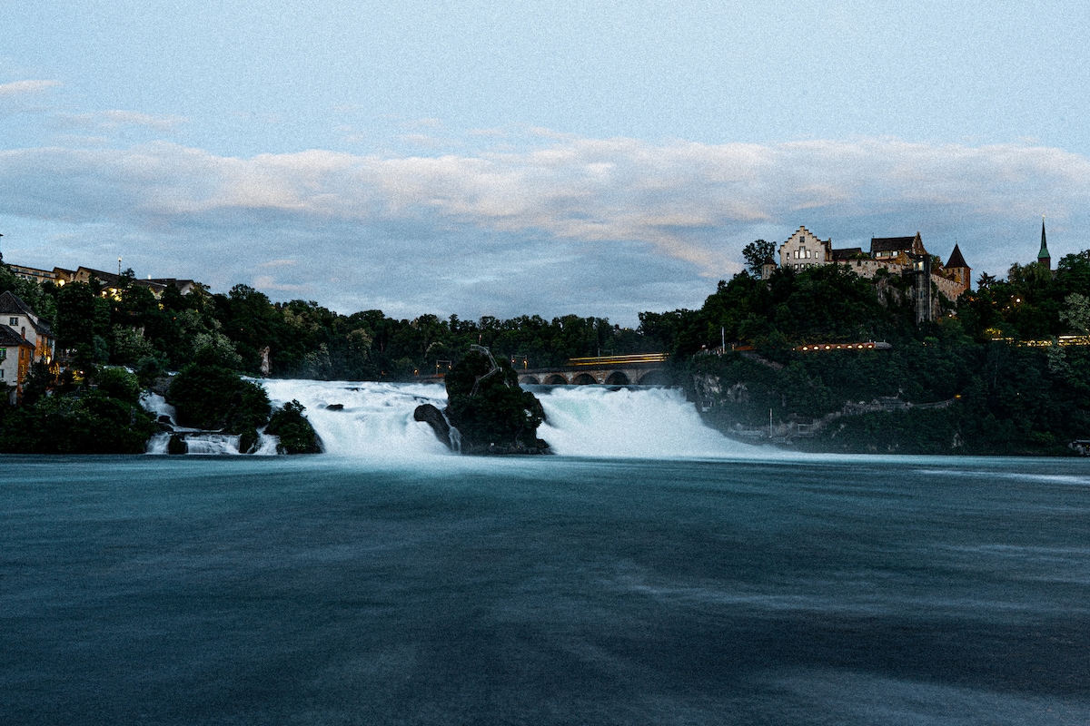 The image captures the impressive flow of the Rhine Falls, with cascading water framed by lush greenery. A nearby hill features architecture, including a mixture of rustic and modern buildings. The tranquil blue water contrasts with the vibrant natural landscape.