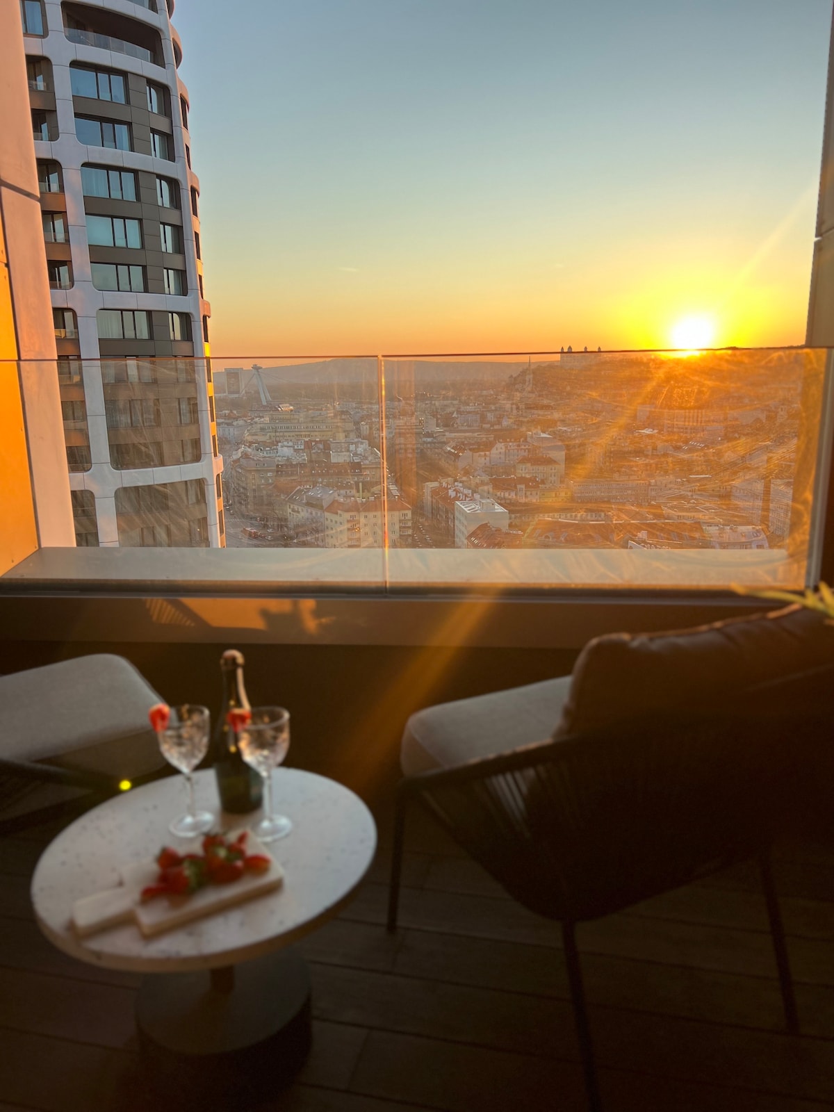 A balcony offers a view of a sunset over the city skyline. The scene includes a small table set with two glasses and a bottle, accompanied by fresh strawberries. Comfortable chairs are positioned invitingly, enhancing the relaxing atmosphere of the space.