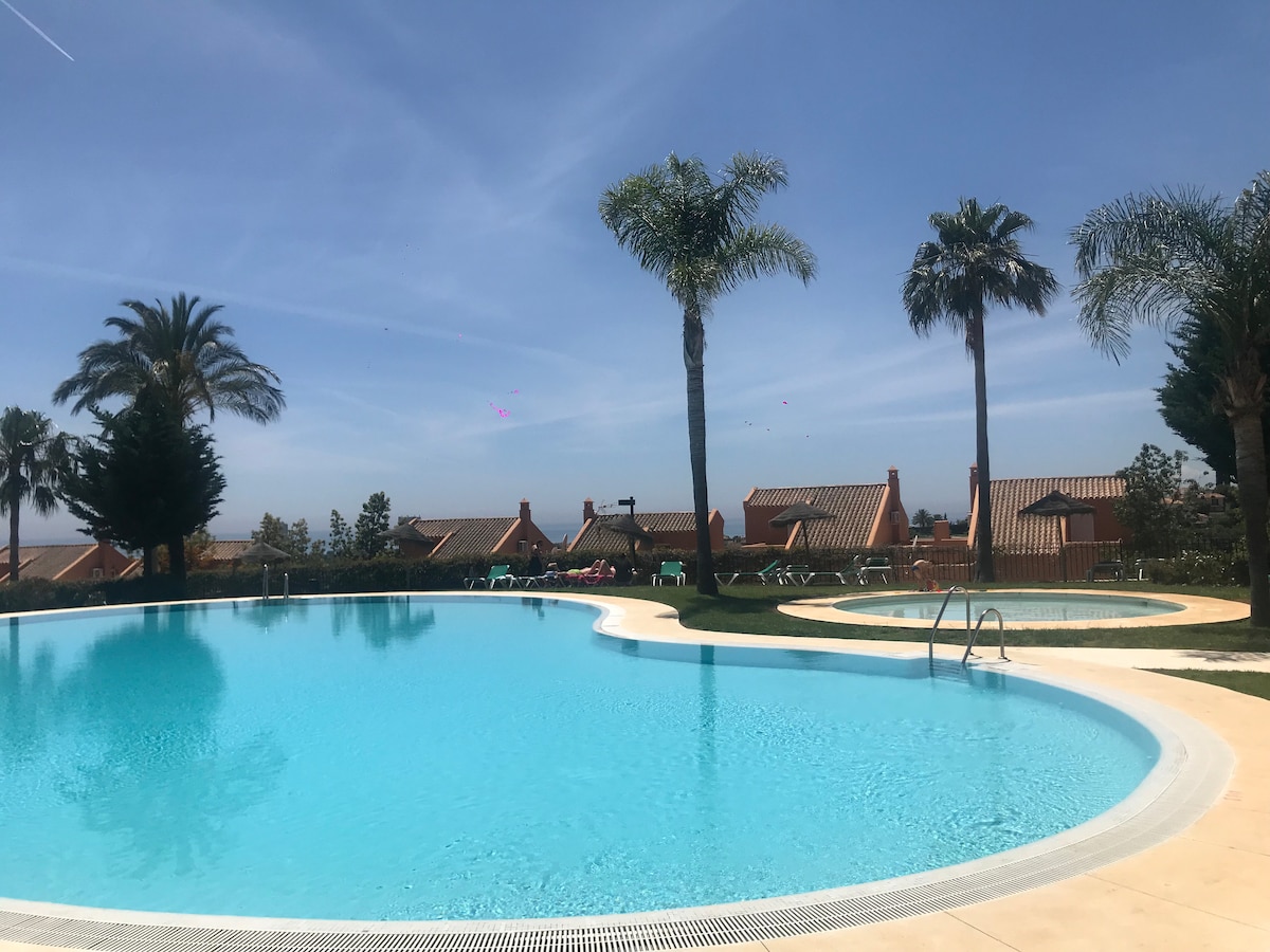 A tranquil pool area is featured, surrounded by lush palm trees and charming villas. The large, clear swimming pool reflects the sky, complemented by a smaller hot tub nearby. Comfortable lounge chairs are arranged around the pool, inviting relaxation under the sun.