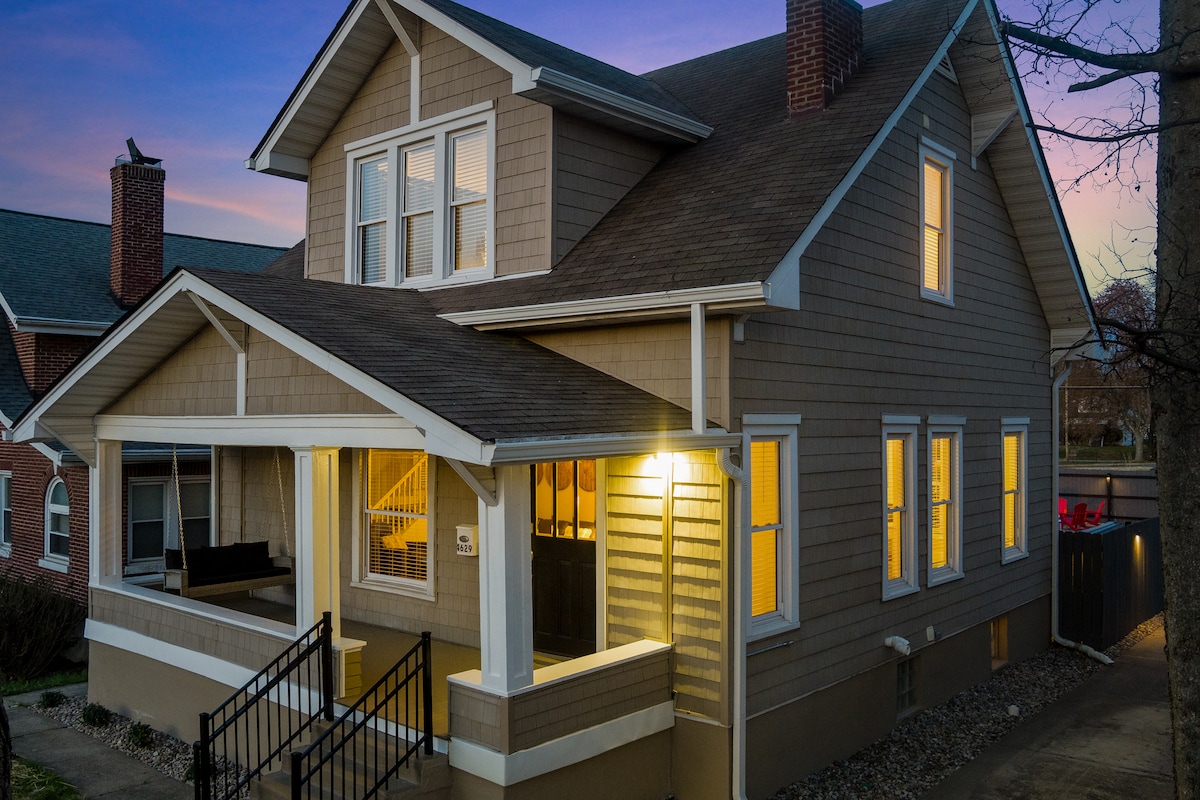 The exterior of the home is depicted at dusk, showcasing a warm glow from the porch lights. The two-story structure features light brown siding, white trim, and multiple windows, creating an inviting appearance. The covered porch offers additional seating, and a stairway leads to the entrance.
