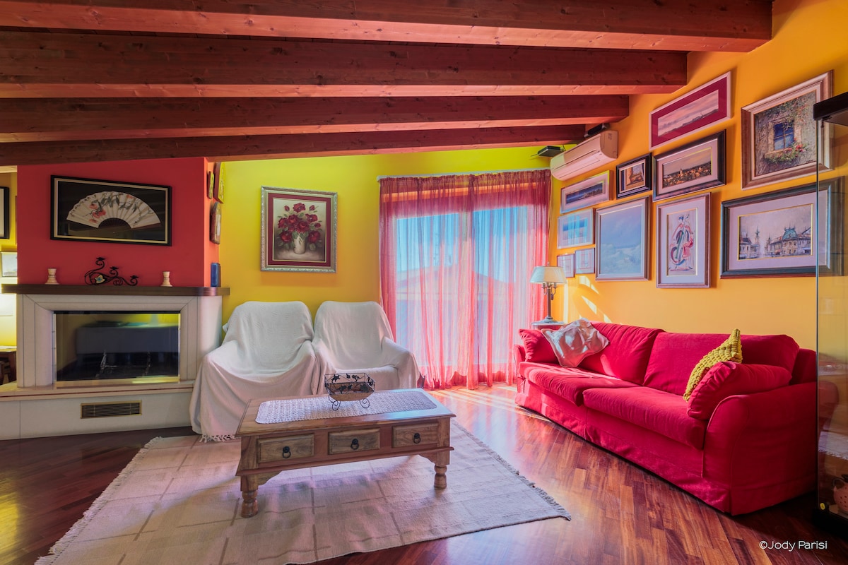 The living area showcases vibrant yellow and red walls, complemented by artwork and framed photographs. A red sofa is positioned centrally, while two white armchairs are draped with covers. Natural light enters through large windows, enhancing the warm ambiance of the space.
