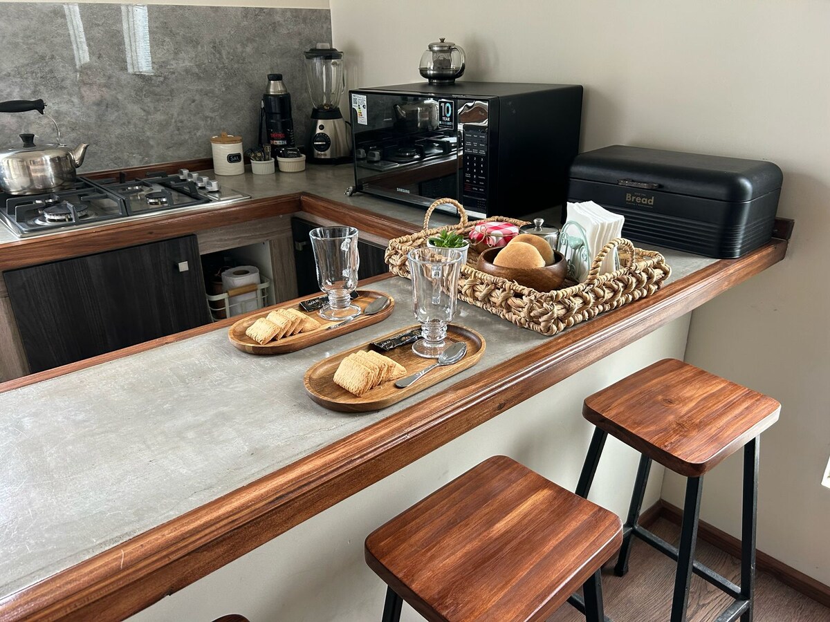 A kitchen countertop displays two wooden trays with snacks, including crackers and bread. A coffee machine, microwave, and various kitchen appliances are present in the background. Four high stools are positioned at the countertop, offering a casual dining area for guests.