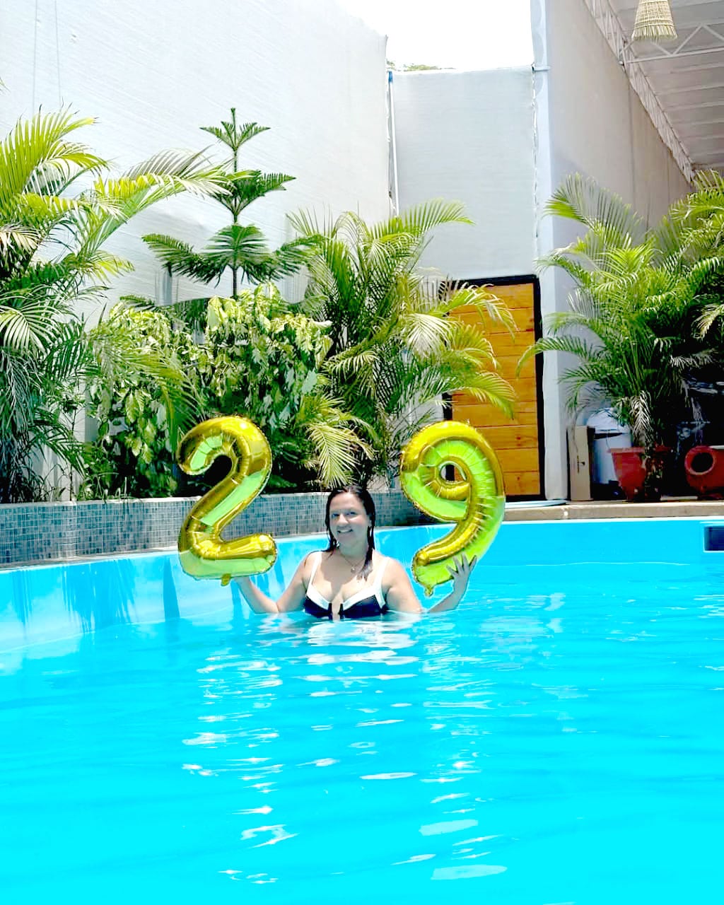 The image captures a vibrant pool scene with clear blue water. Two golden balloons displaying the numbers '2' and '9' are held above the water’s surface. Lush green plants surround the pool, enhancing the tranquil atmosphere.