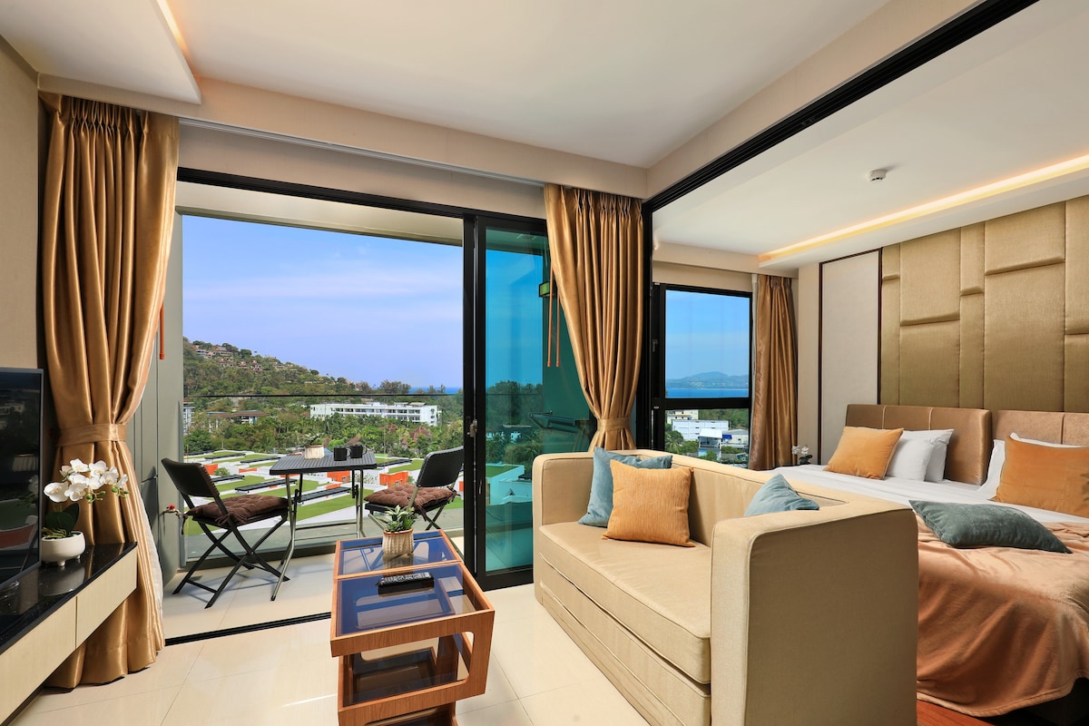 A spacious room is presented, featuring a large bed and a comfortable sofa. Floor-to-ceiling windows offer a view of the outdoors, with a small balcony that includes two chairs and a table. Soft curtains frame the windows, enhancing the bright ambiance.