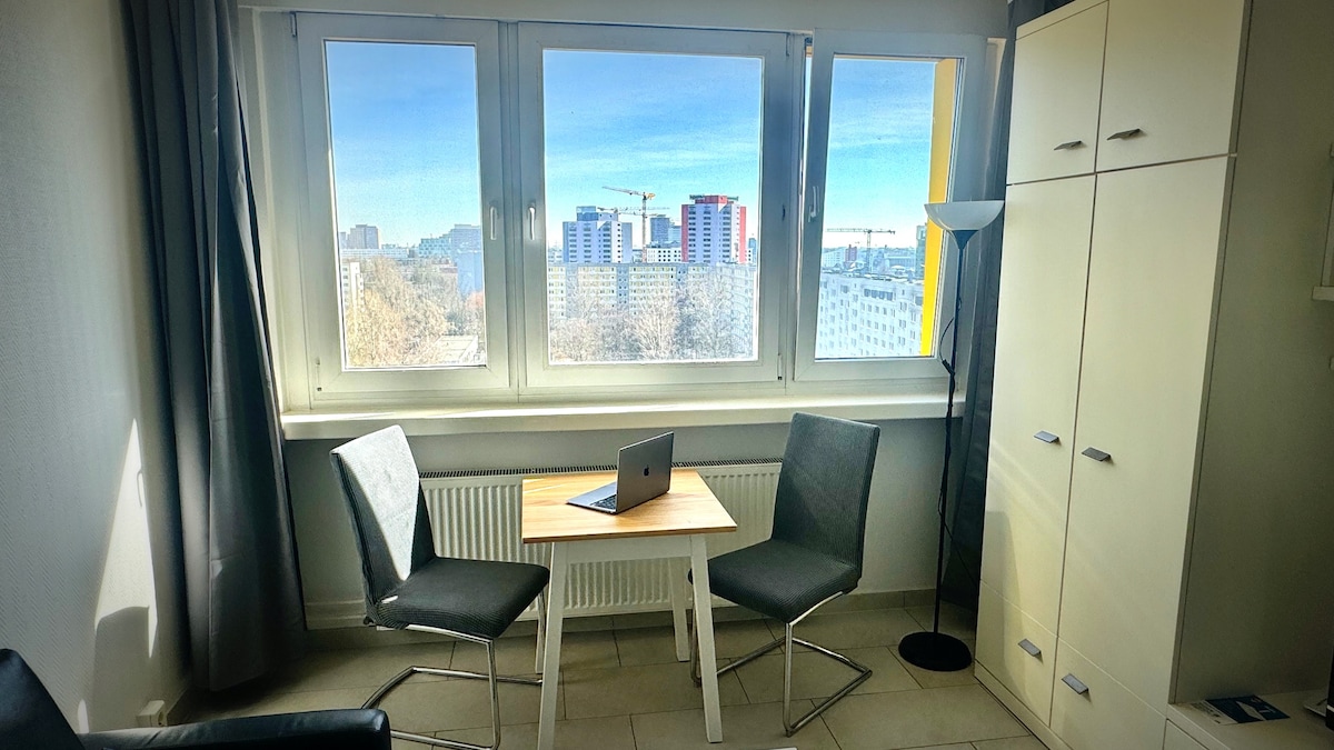 A bright living area features two chairs positioned around a wooden table, complemented by a laptop set atop it. Large windows provide a view of the urban landscape, while a tall white storage cabinet stands nearby, contributing to the clean and modern aesthetic.