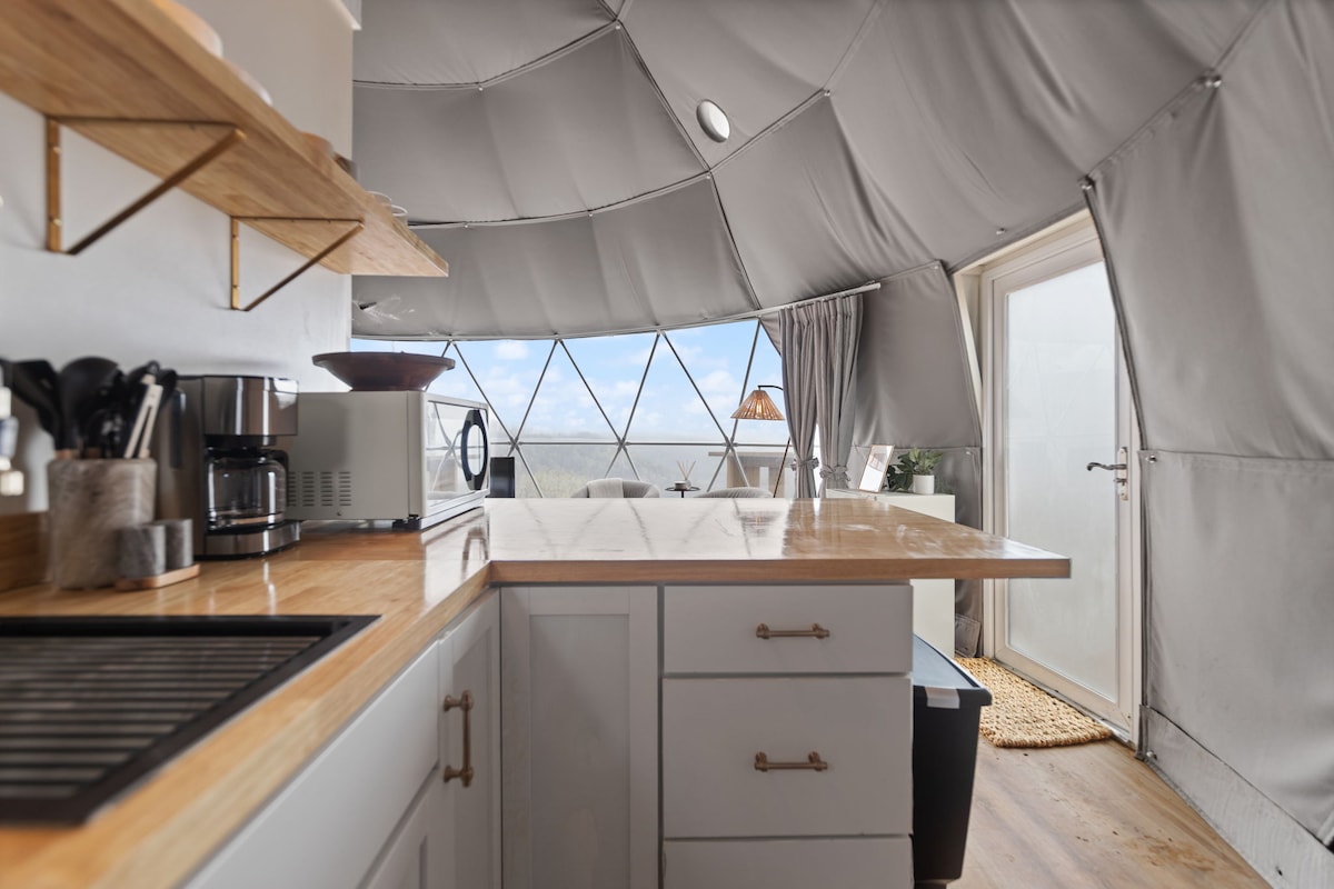 The kitchen area showcases a light wooden countertop with modern appliances, including a coffee maker and microwave. Natural light floods through expansive windows, which enhance the airy feeling. Sleek shelves are mounted on the walls, holding kitchen essentials for convenience.