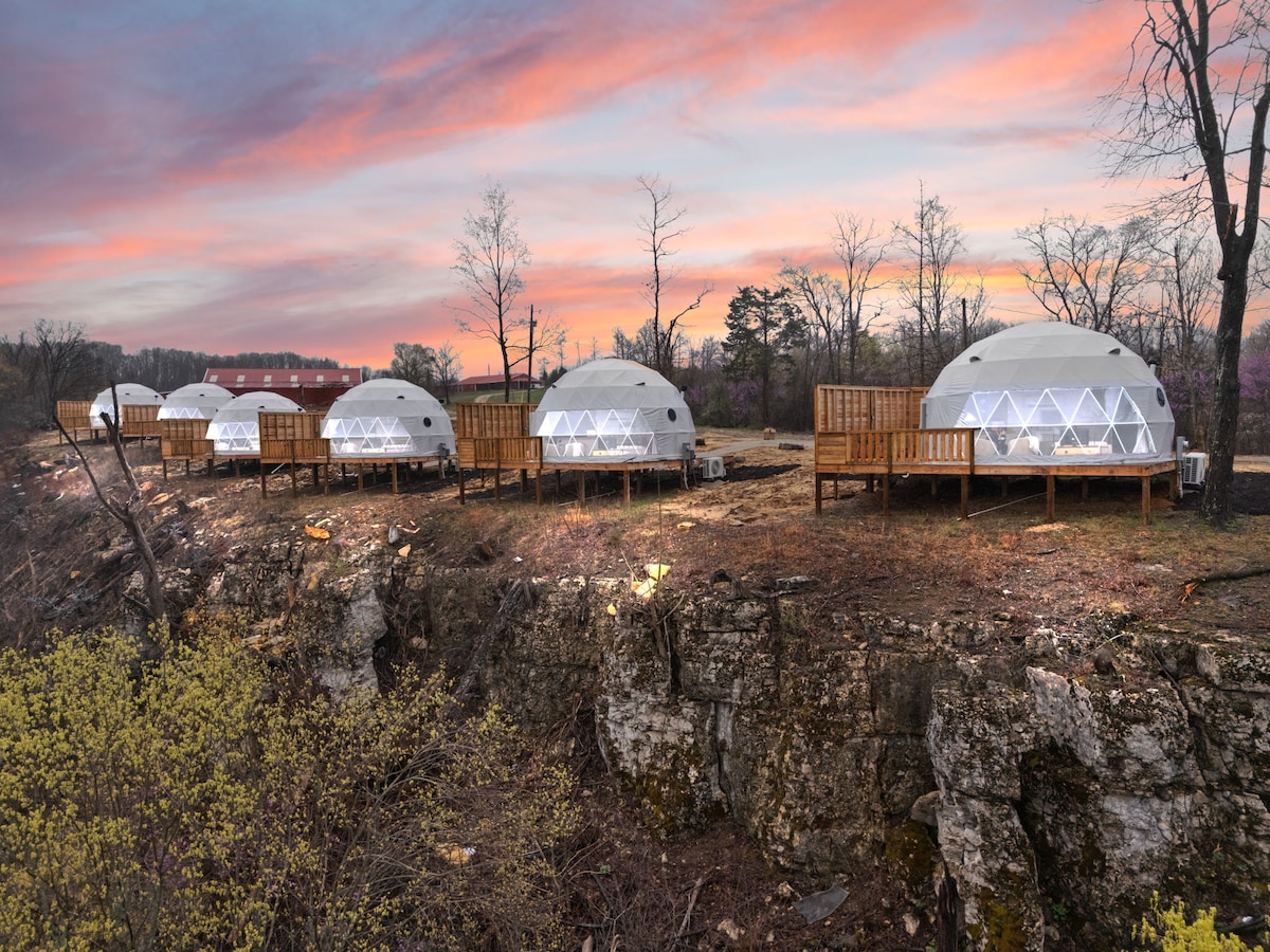 A row of six geodesic domes is situated on a ridge, surrounded by natural foliage. Each dome features large, triangular windows and is elevated on wooden decks. The soft colors of a sunset enhance the serene landscape, creating a peaceful atmosphere.