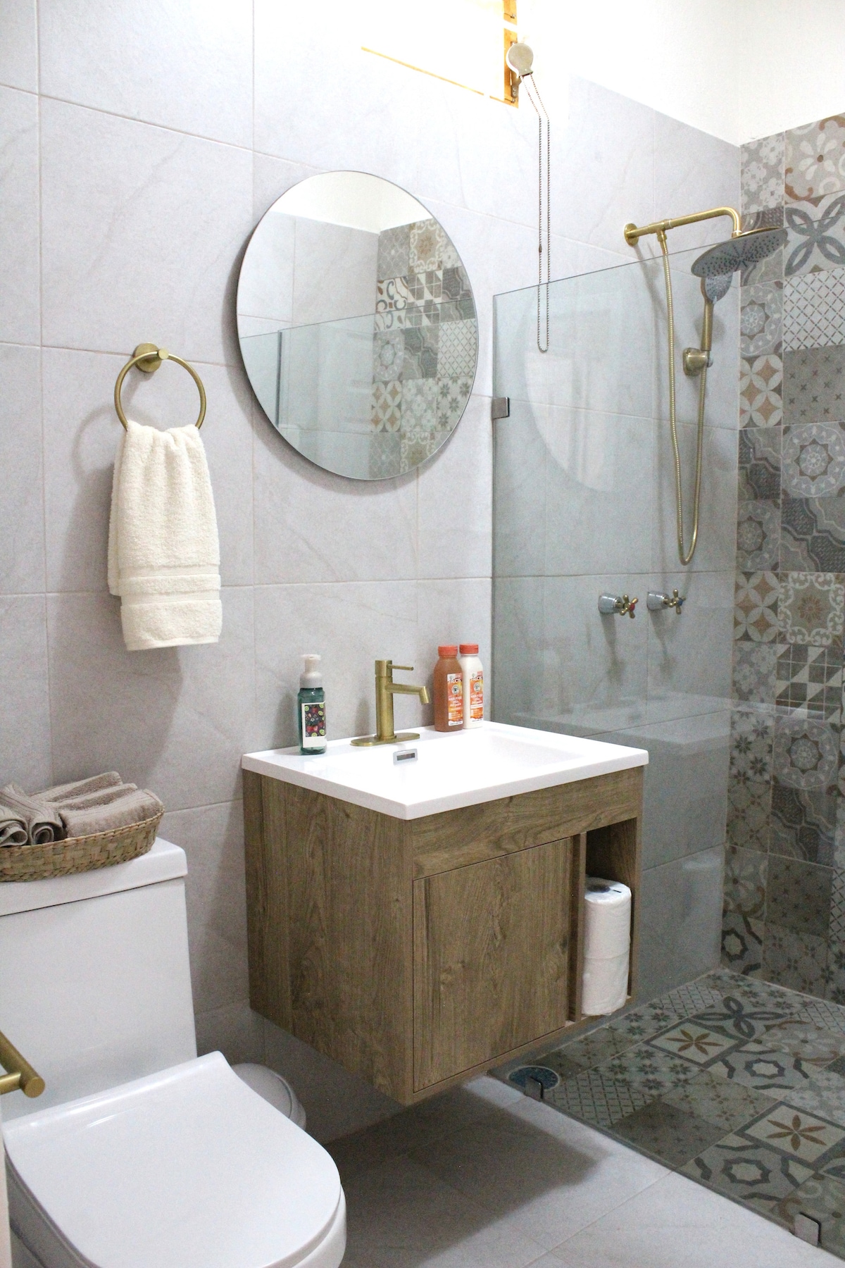A modern bathroom features a compact vanity cabinet with a white sink. A circular mirror hangs above, and a walk-in shower is enclosed in glass, displaying textured tiles. Soft towels are neatly arranged on a towel ring, contributing to the functional design.