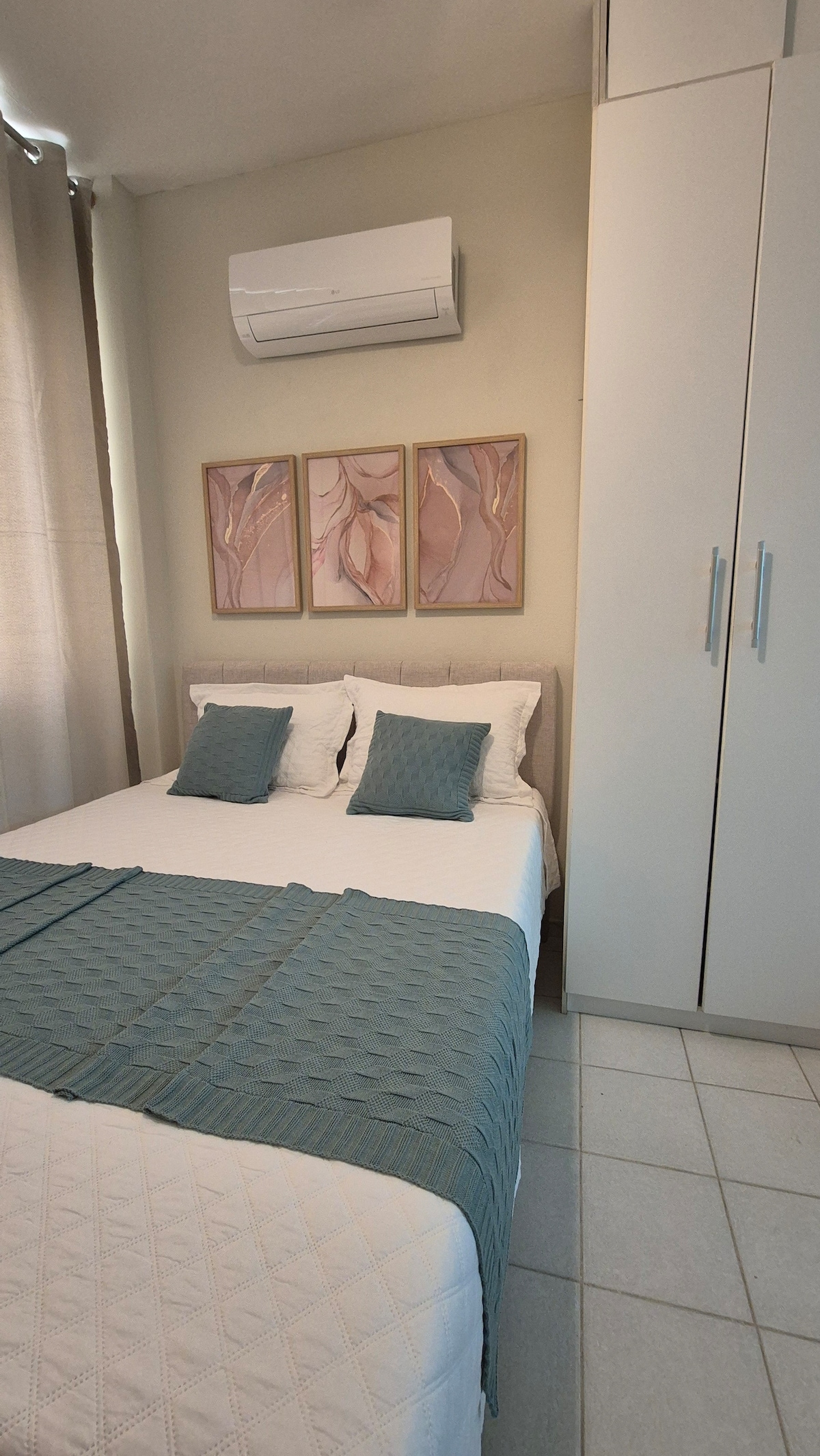 A calming bedroom setting is presented, featuring a spacious bed with a textured blue throw draped over it. Soft pillows are arranged neatly, and three art pieces adorn the wall above the bed. A sleek white wardrobe is positioned nearby, and a window allows natural light to filter in.