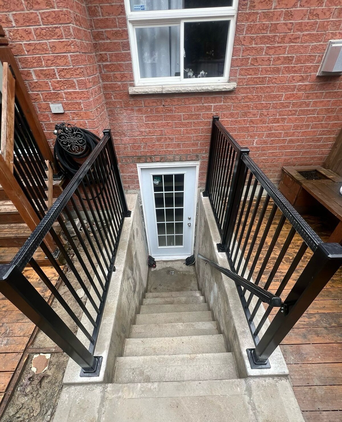 A set of concrete stairs is visible, leading down from a wooden deck to a white door with glass panels. Black metal railings flank both sides of the staircase, enhancing safety and accessibility. The brick wall is prominently featured in the background.