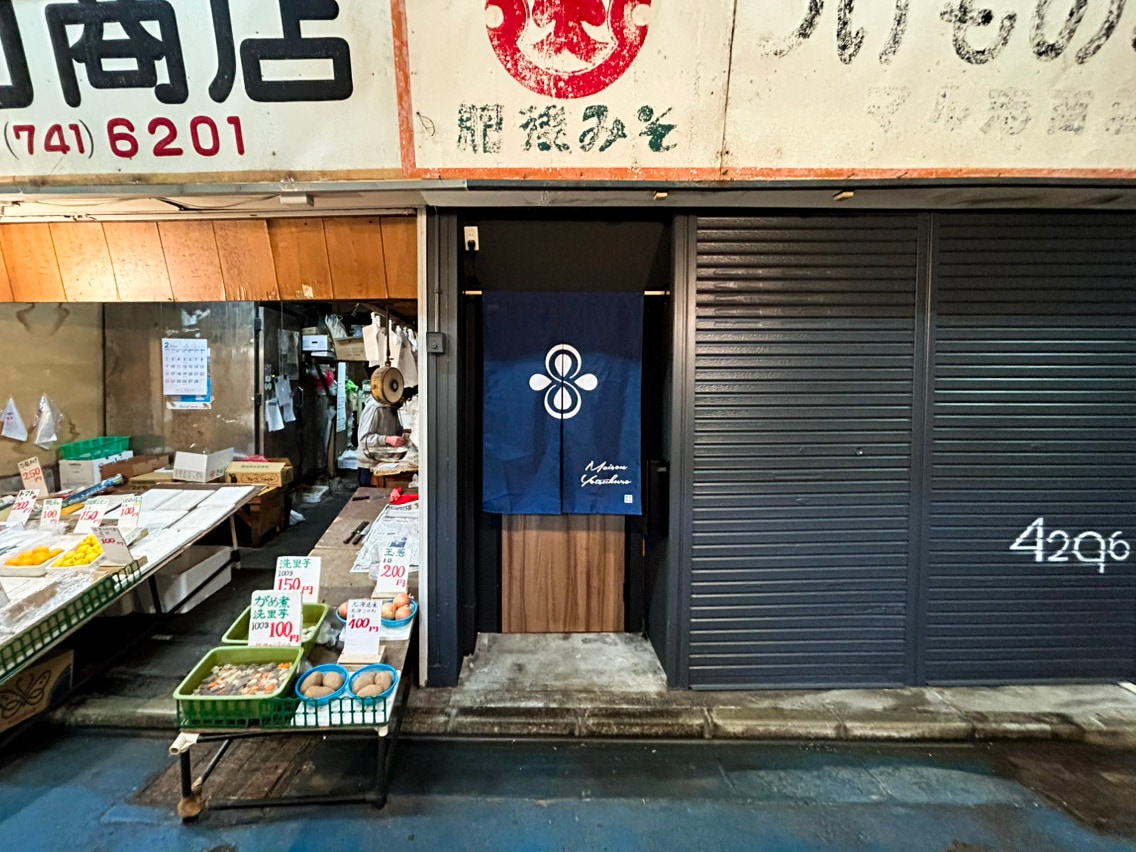 A market entrance is displayed, featuring a blue curtain adorned with a stylized logo. Stall displays with fresh produce, packaged goods, and price signs line the walkway, contributing to a lively marketplace environment. The storefronts showcase a mix of traditional and contemporary elements.