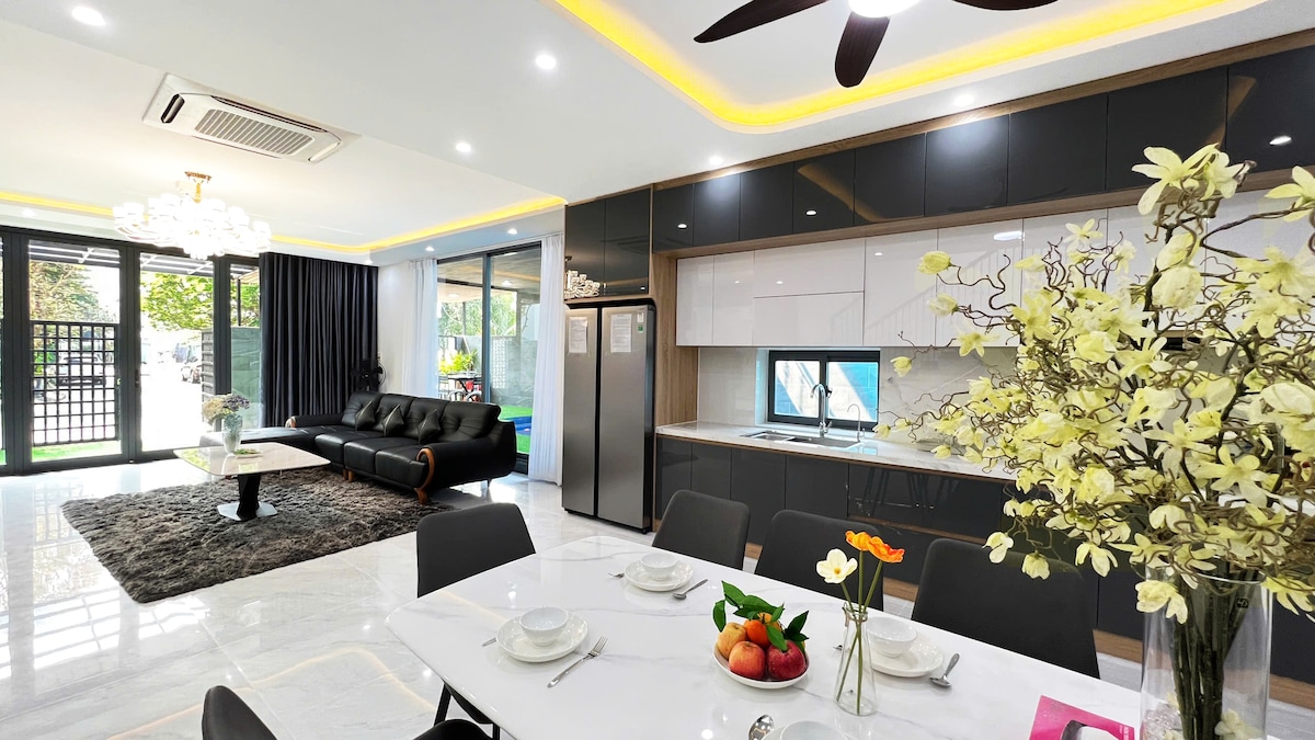 A modern kitchen and dining area is presented, featuring sleek cabinetry and a marble dining table set for six. A comfortable black sofa is positioned near large glass doors that open to a vibrant outdoor space, complemented by decorative flowers and fruits arranged on the table.